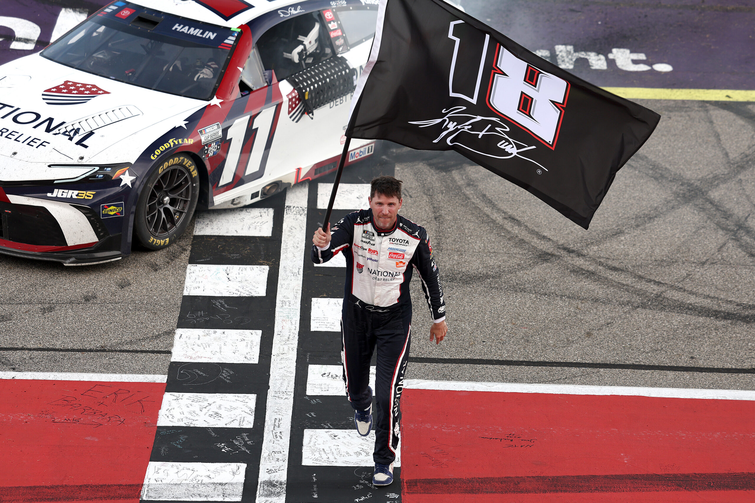 BROOKLYN, MICHIGAN - JUNE 07: Denny Hamlin, driver of the #11 National Debt Relief Toyota, waves an 18 - 8 flag in honor of the late Kyle Busch after winning the NASCAR Cup Series FireKeepers Casino 400 at Michigan International Speedway on June 07, 2026 in Brooklyn, Michigan.